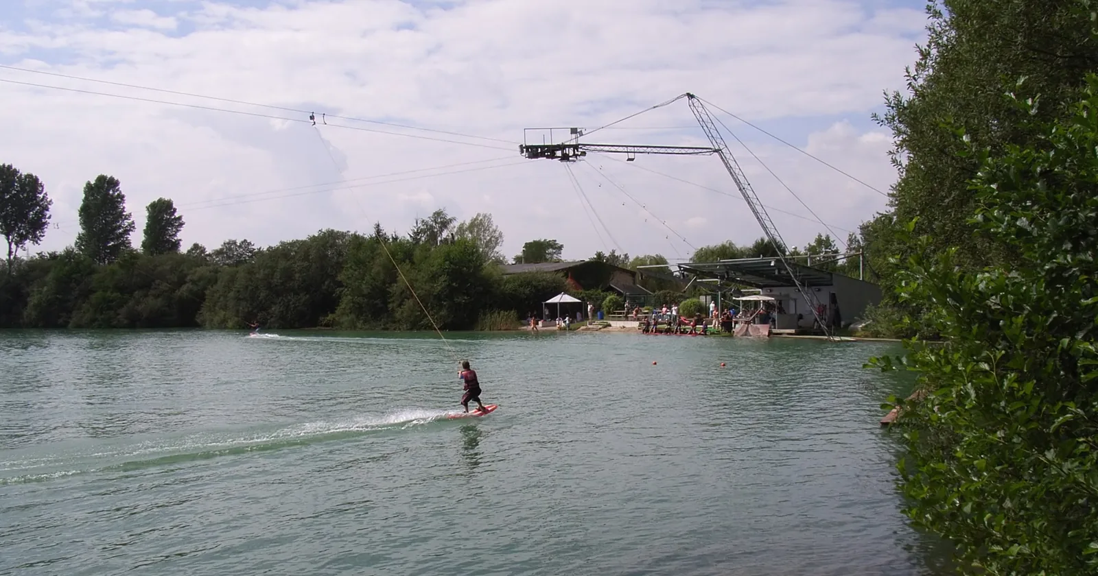 Wakeboarder towed by a full-size overhead cable past the corner tower and carrier at a cable park lake, the system Al Forsan runs in Abu Dhabi