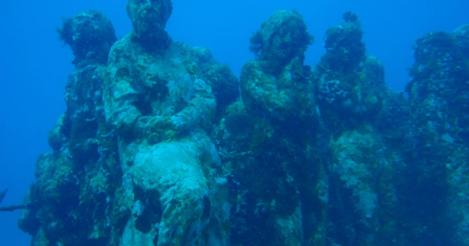 Life-size figures of MUSA, the underwater sculpture museum between Cancun and Isla Mujeres, overgrown with algae and coral in blue water, a stop on most Cancun diving trips