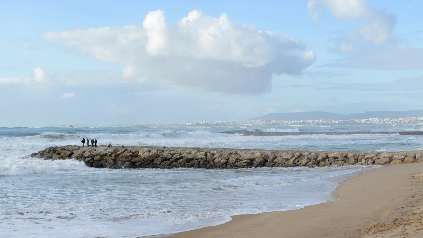 Heavy swell breaking over a rock groyne at Costa da Caparica, Portugal, with Lisbon's coastline across the water