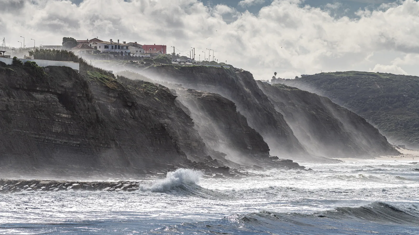 A wave breaking below the cliffs at Ericeira, Portugal, with spray blowing back off the face and the village on the clifftop