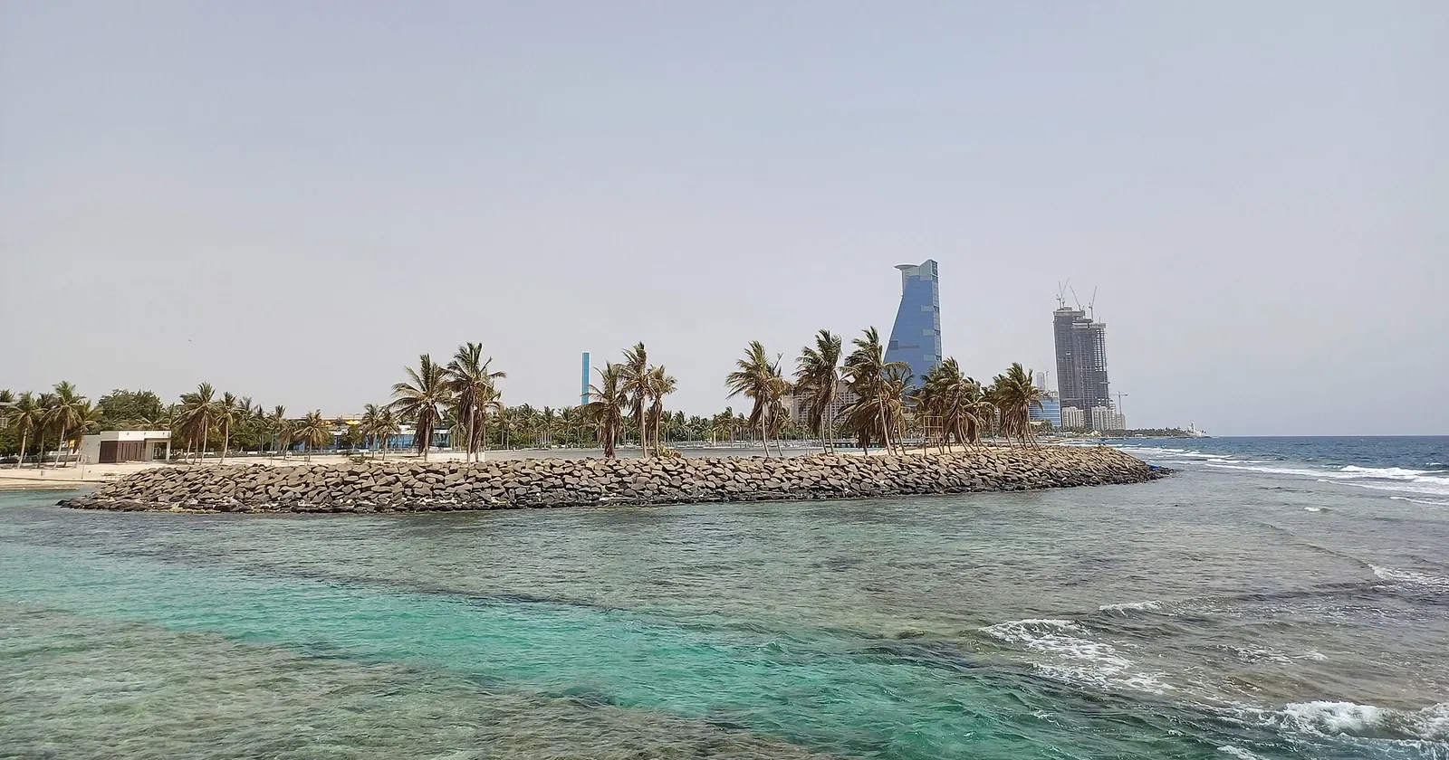The shallow fringing reef off the Jeddah seafront, with a palm-lined rock breakwater and the city's towers behind, the coast Jeddah diving starts from
