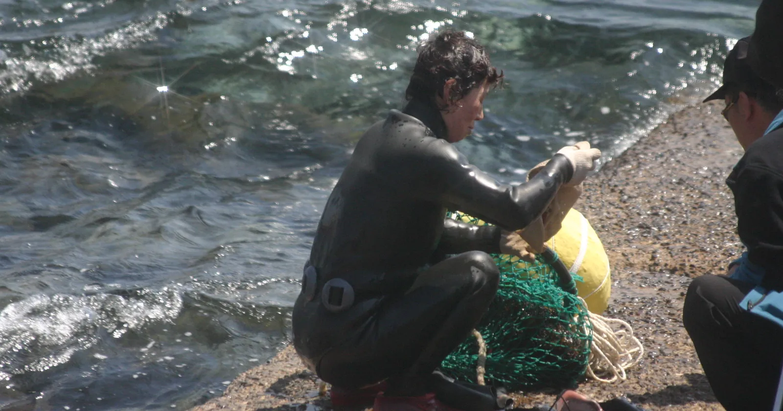 A Jeju haenyeo in a black wetsuit sorting her net bag beside a float on the rocks at the water's edge, the free-diving tradition that shapes the rules for Jeju diving
