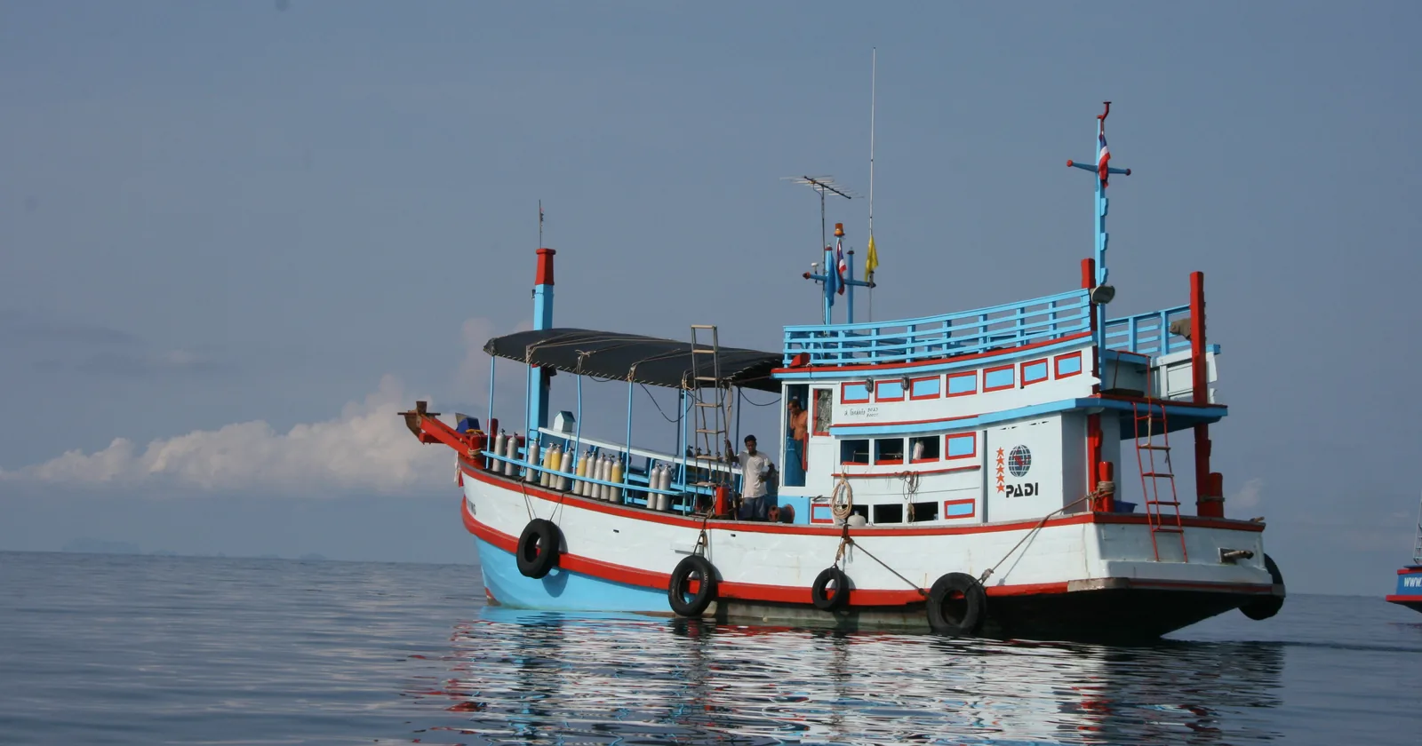 Thai wooden dive boat moored off Koh Tao with scuba cylinders racked along the rail and a PADI centre board on the wheelhouse