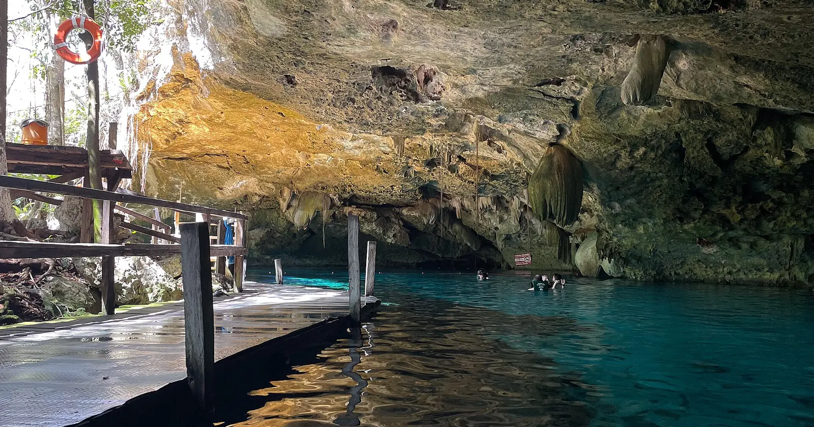 The daylight zone of Cenote Dos Ojos on the Riviera Maya, with a wooden platform, stalactites and snorkellers in clear water