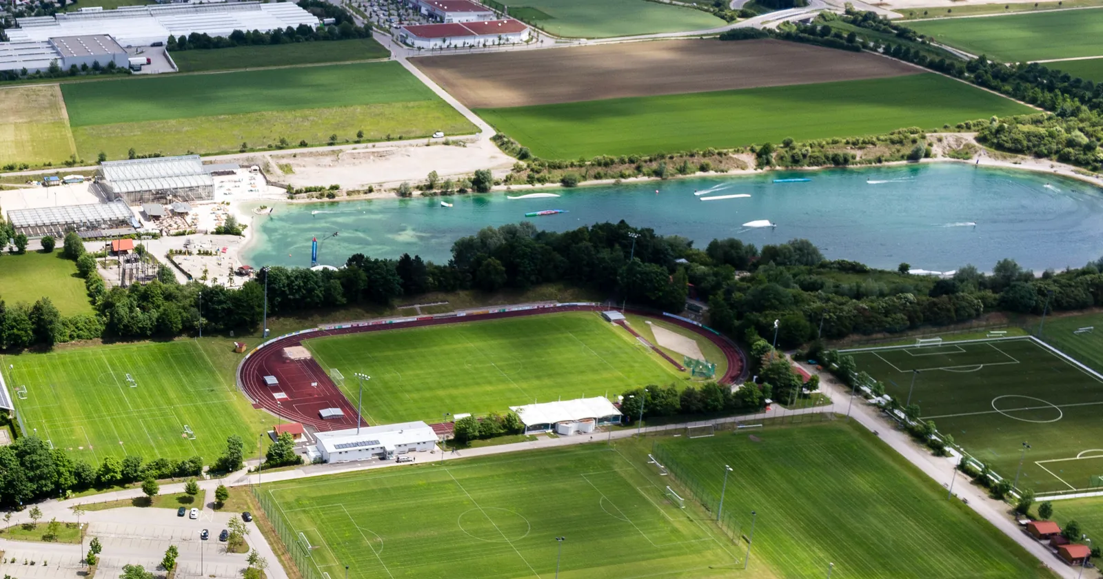 An aerial view of Sportpark Aschheim near Munich: a turquoise lake with floating wakeboard obstacles, a sand beach, and football pitches and an athletics track beside it
