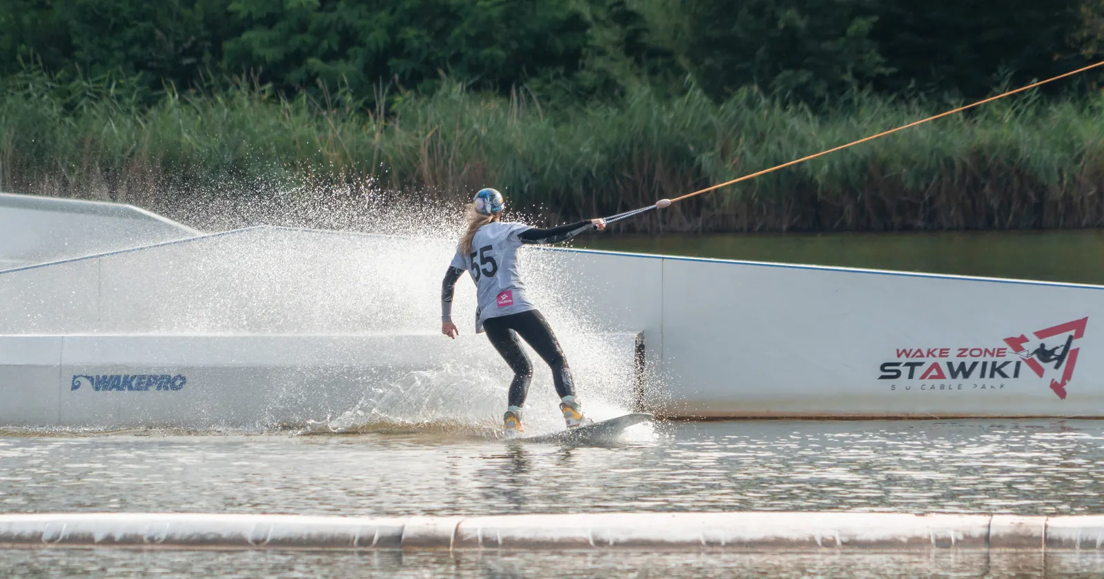 A wakeboarder pulled by an orange cable line past a slider at a Polish cable park, throwing spray behind the board